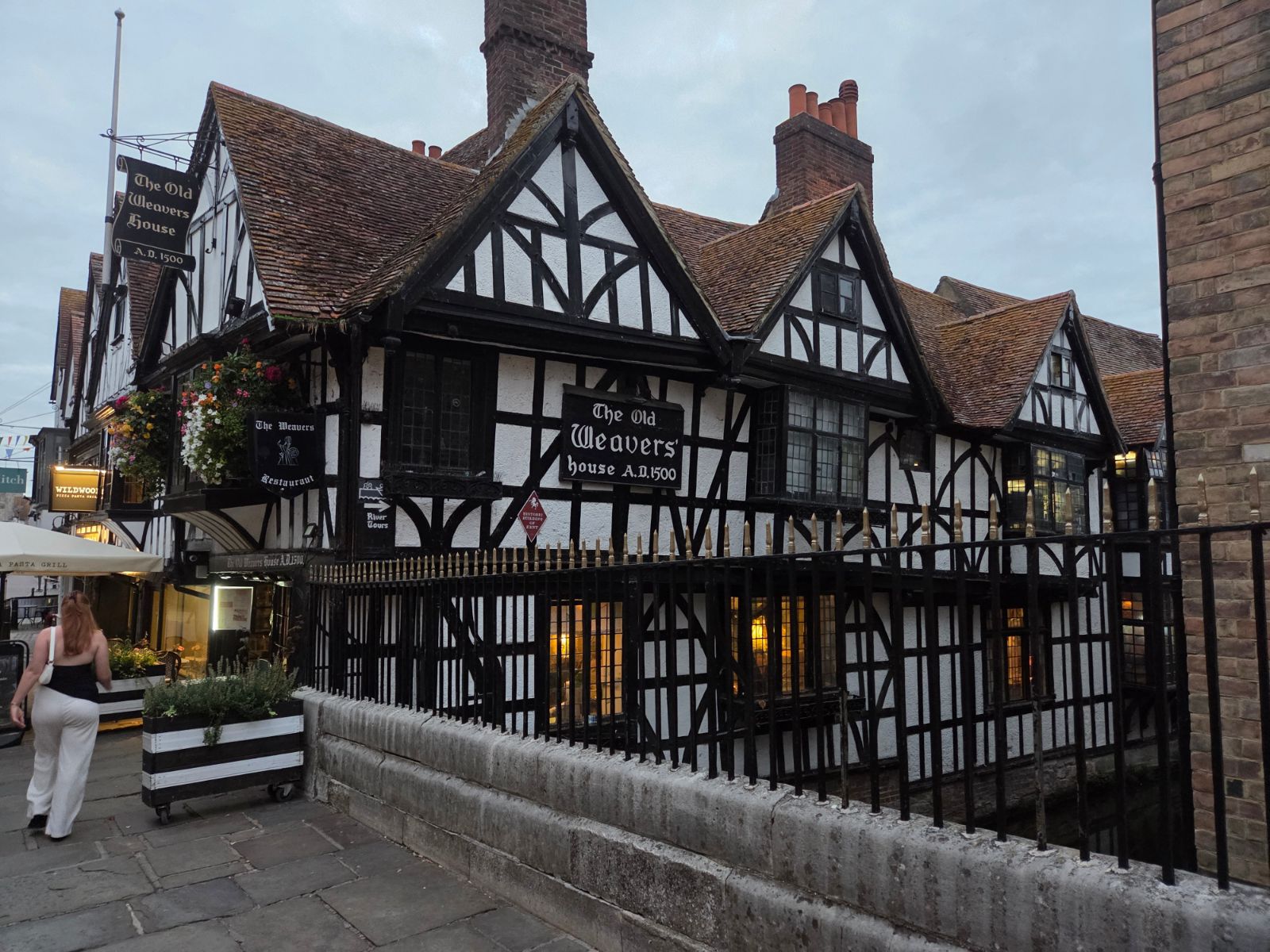 Black and white timber-framed Old Weavers' House (A.D. 1500) in Canterbury at dusk, with hanging flower baskets and iron railings along the river